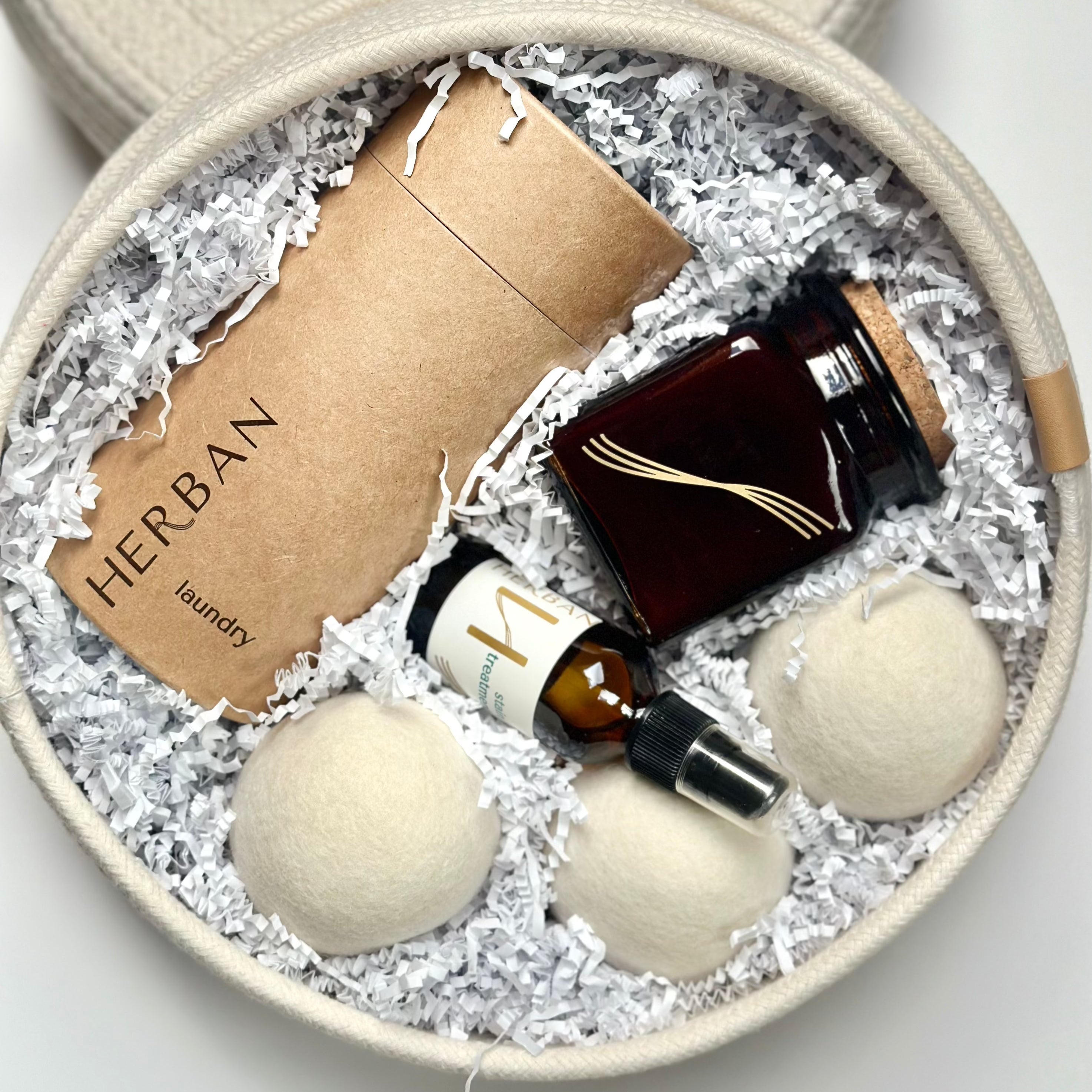 Cotton round rope basket filled with Laundry powder packaged in a paper tube, Wool dryer balls, soy candle in square amber glass and an amber glass of stain removing spray.