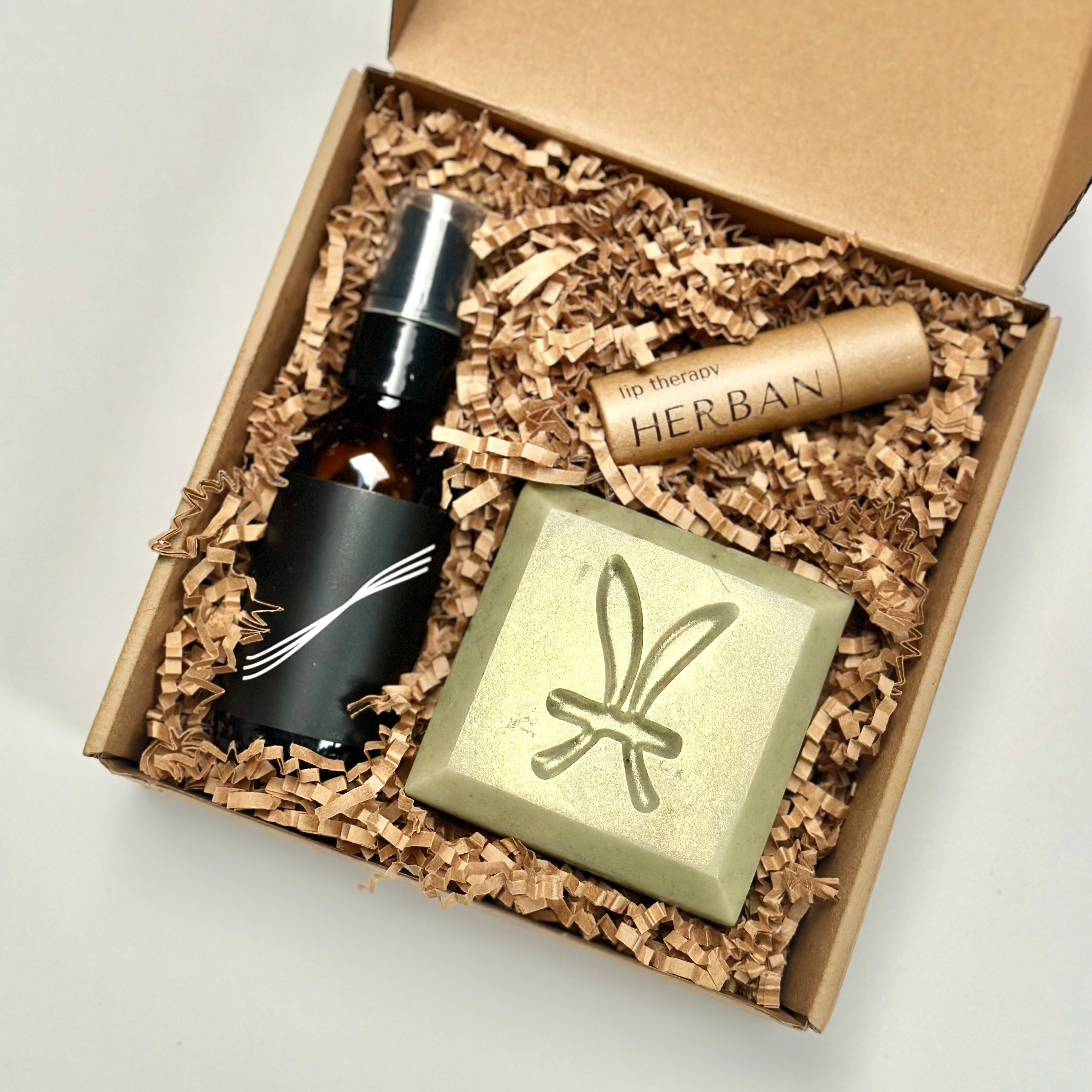 A flat kraft tuck top box filled with a 2oz amber glass hand sanitizer gel bottle with pump, a green square and beveled edge soap bar and a paper tube packaged lip balm