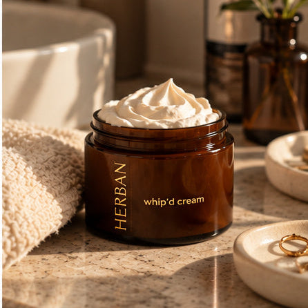Open jar of Whip'd Cream with a swirl of white moisturizer on a bathroom counter beside a woven towel