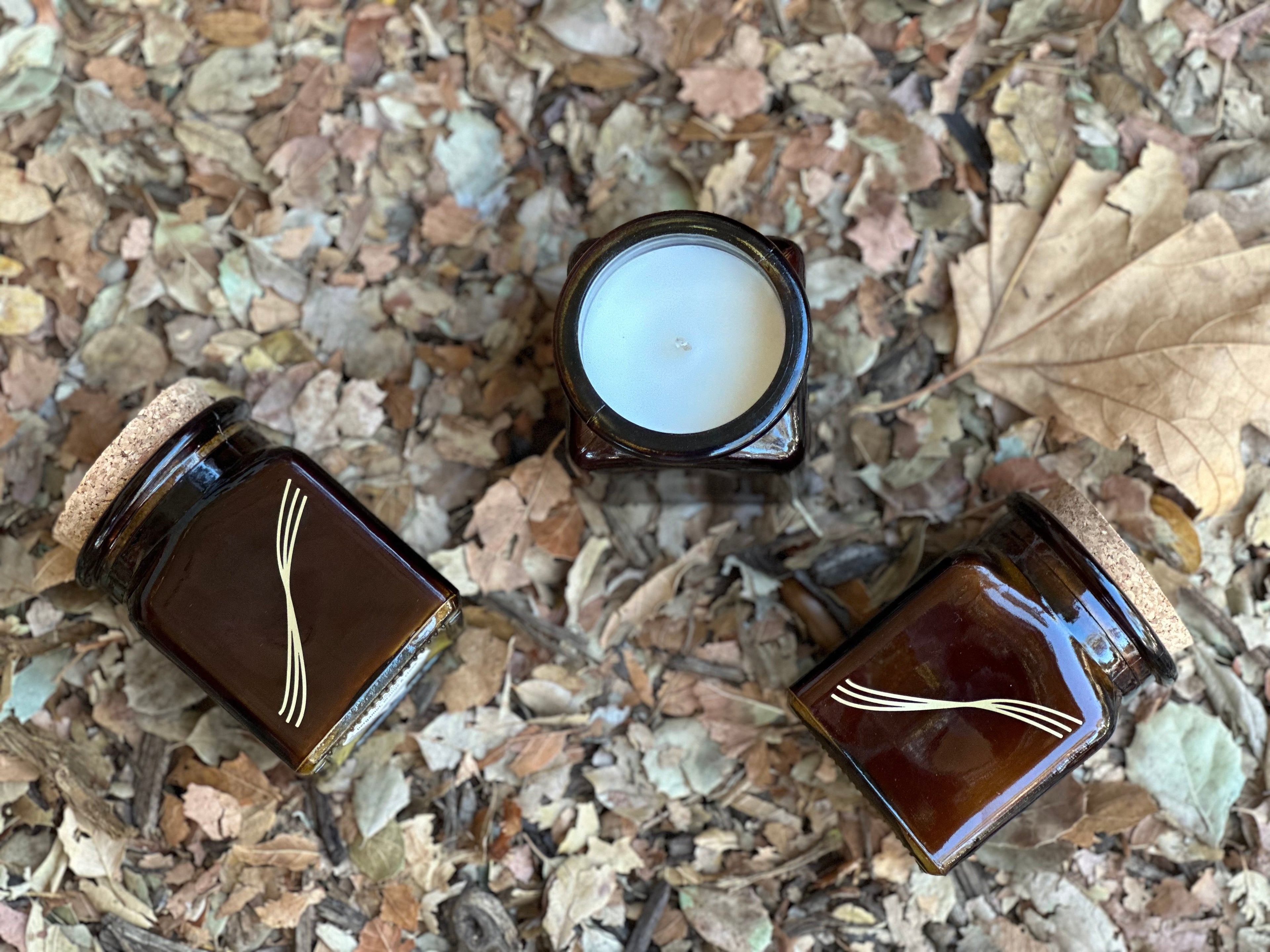 3 square amber glass candle jars with metallic gold printed symbol, laying amongst golden fallen leaves
