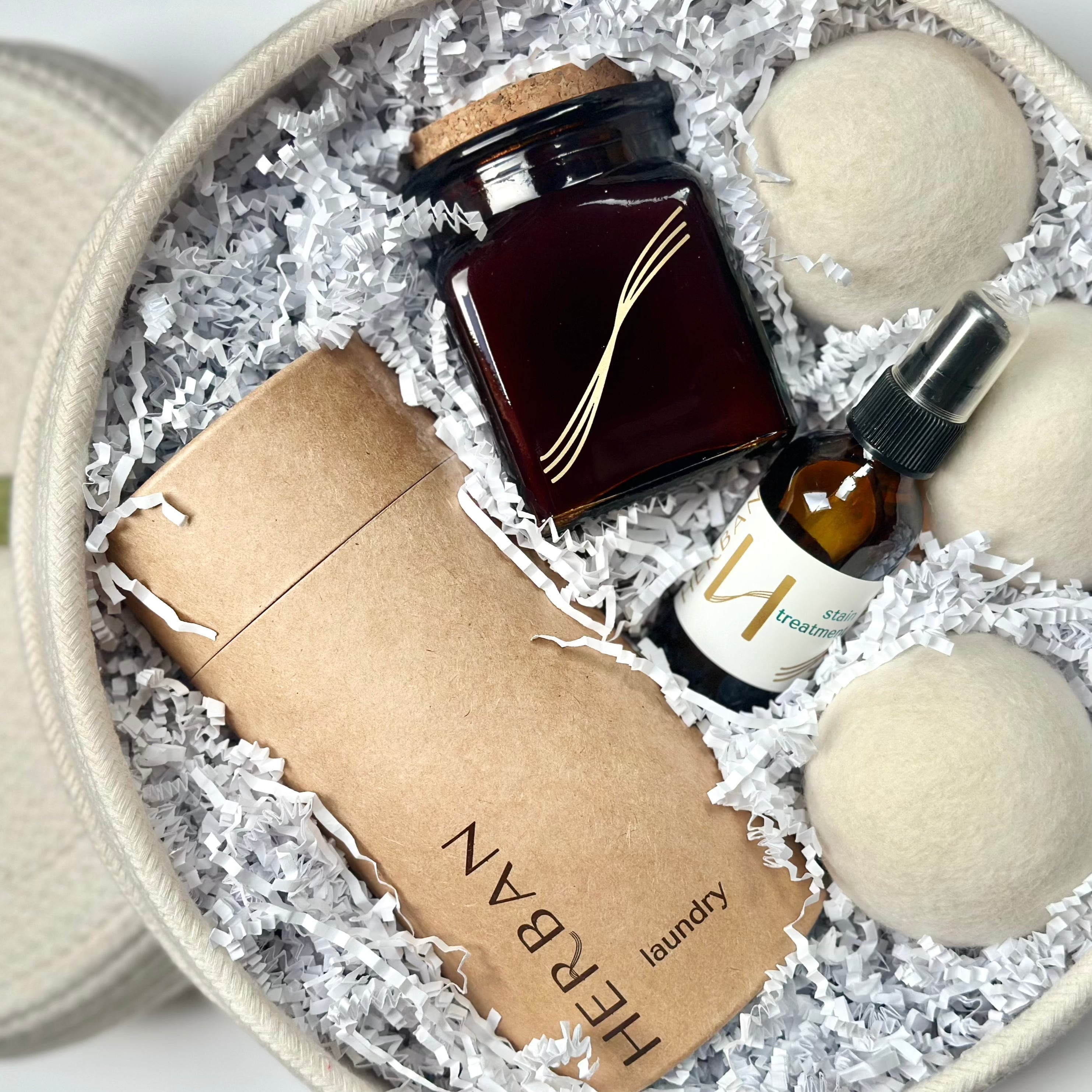 Cotton round rope basket filled with Laundry powder packaged in a paper tube, Wool dryer balls, soy candle in square amber glass and an amber glass of stain removing spray.  