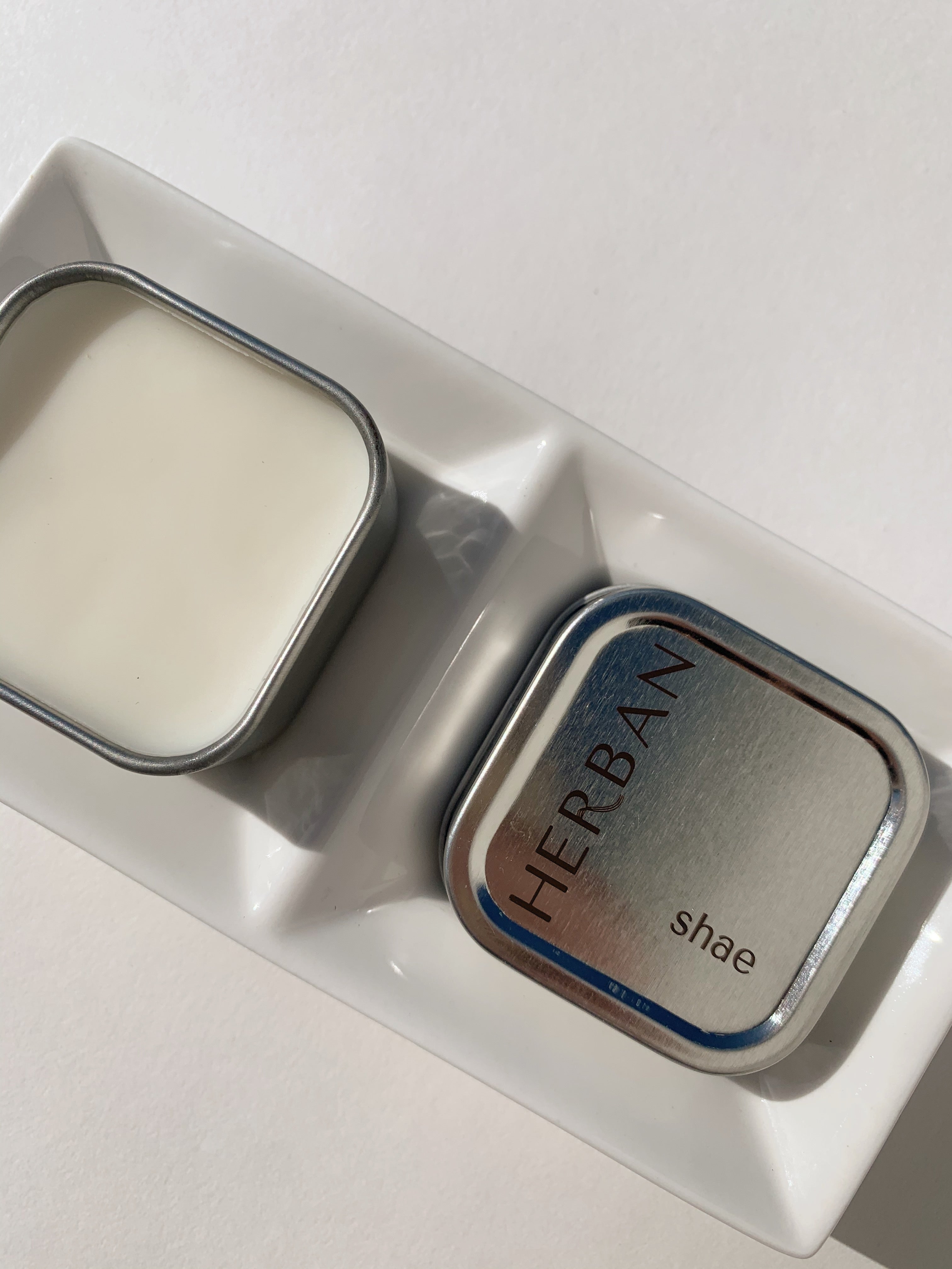 Herban Body Care's shea butter balm in a square silver tin with the lid off and showing the solid cream colored moisturizer