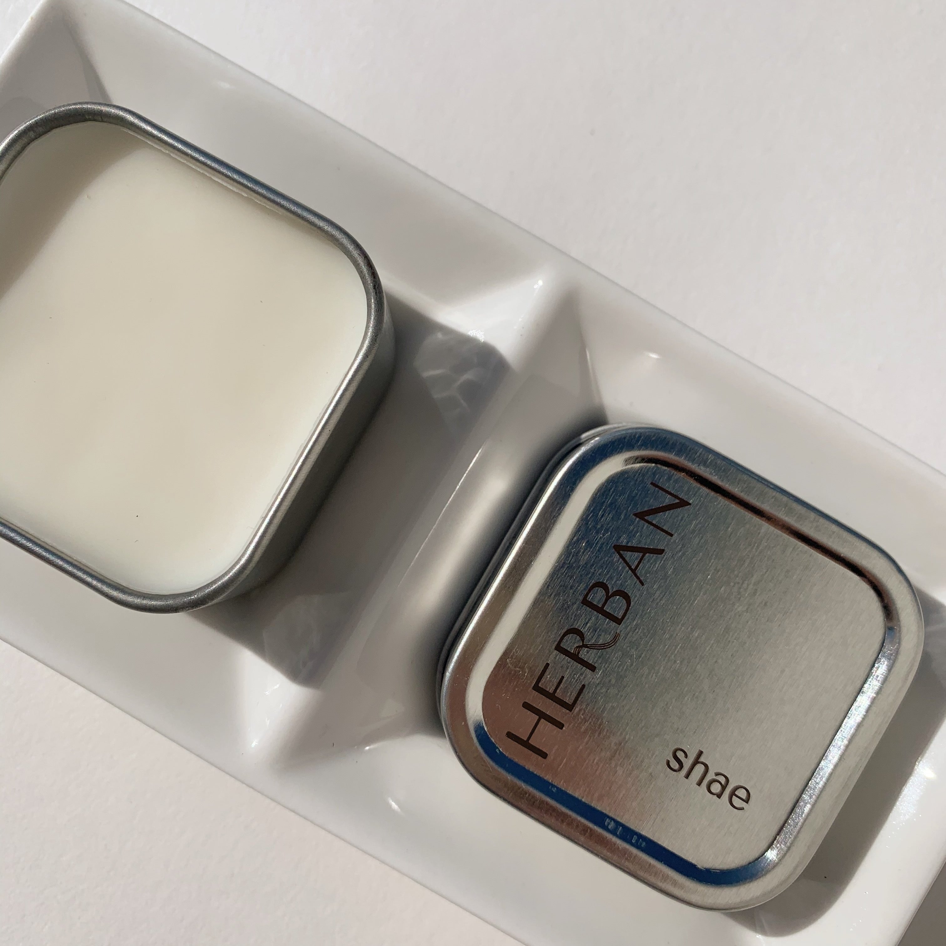 Herban Body Care's shea butter balm in a square silver tin with the lid off and showing the solid cream colored moisturizer